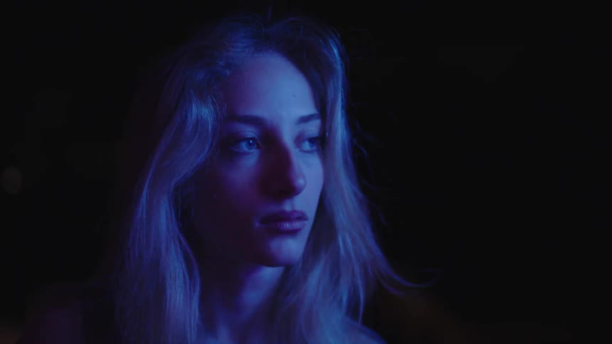 A woman with long light hair is illuminated by soft blue light as she looks sad