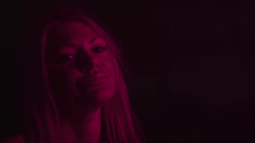 A close view shows a woman with long light colored hair wearing a thin chain necklace and looking off to her right with a serious expression with a red light illuminating the left side of her face