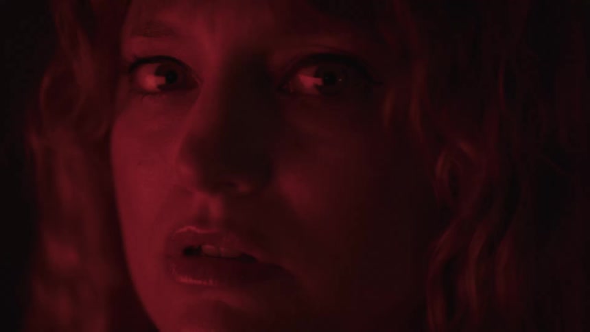 A close view shows a person with long light colored hair and a worried expression lit by a reddish light as they turn their head and look straight into the camera