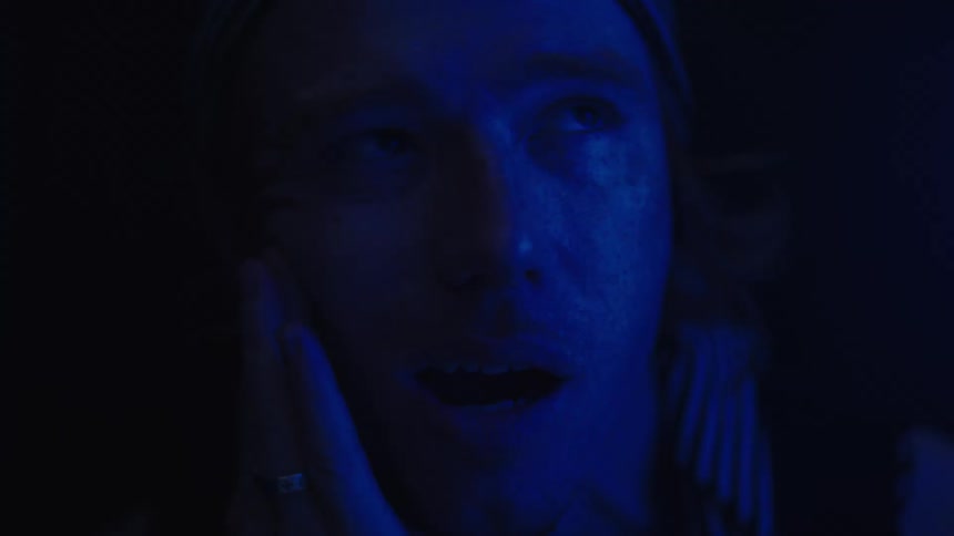 A young man in a dark room dimly illuminated by blue light ties a scarf over his head looking tired