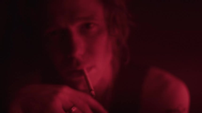 The video shows a man with shoulderlength hair in a dimly lit setting he is holding a cigarette to his lips and exhaling smoke