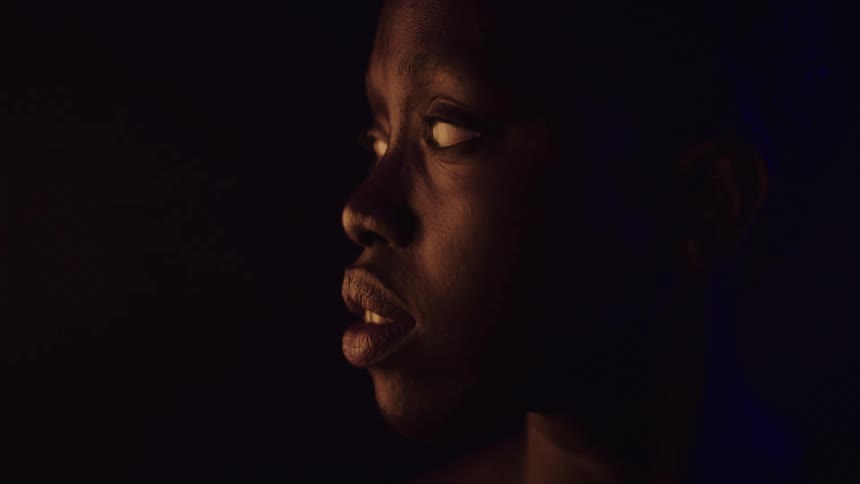A close view shows a woman with dark skin in profile with her mouth slightly open and their head turning from left to right lit from the front with a warm orange glow on the left and a faint blue light on the right