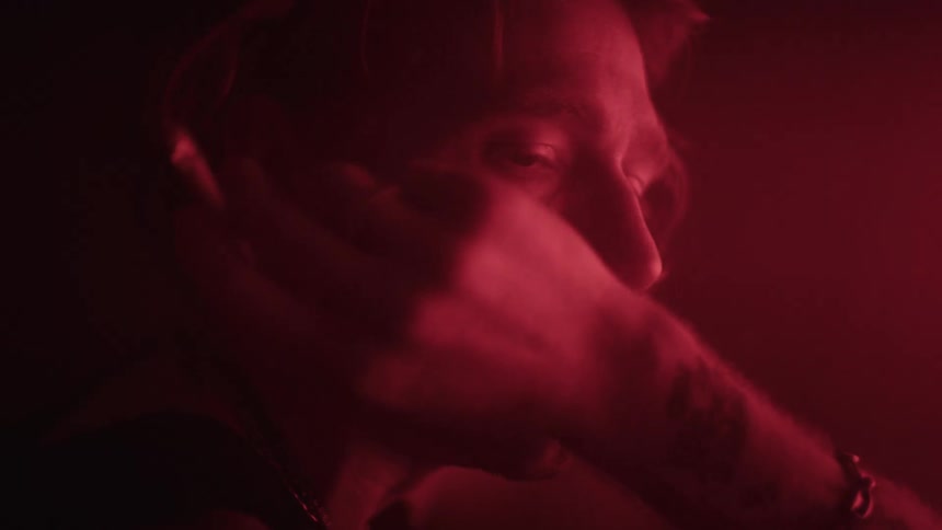 A young man with a tattoo on his arm sits in a dark room illuminated by red light smoking a cigarette