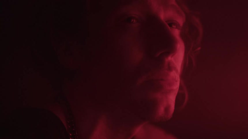 A close view shows a man with long blonde hair and a necklace holding a lit cigarette and looking up as a red light illuminates his face and hair