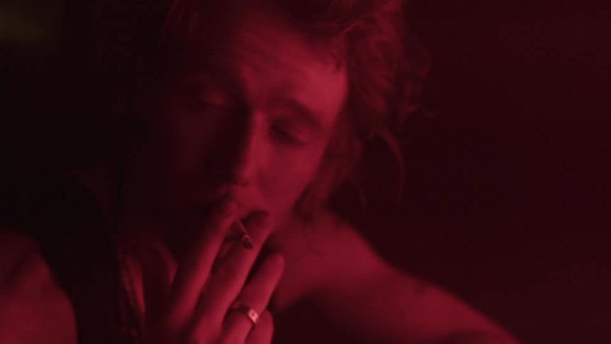 A man with light colored hair and a chain necklace looks up and then down holding a cigarette to his lips as his face is lit by a red light