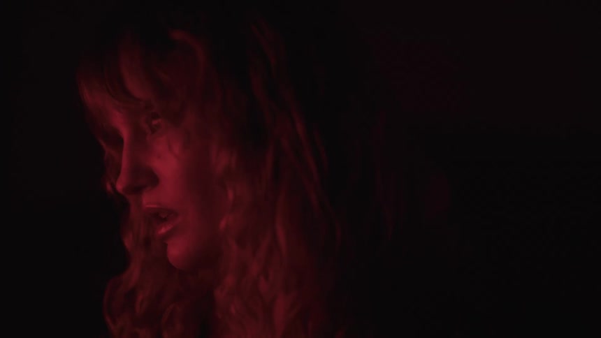 A young blonde woman with long hair stands inside a dark room illuminated by dim red light