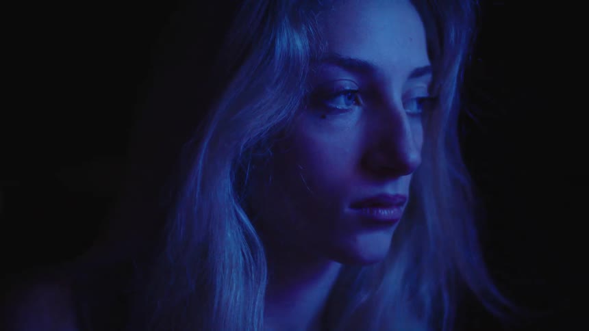 A young woman in blue light stares seriously at the camera