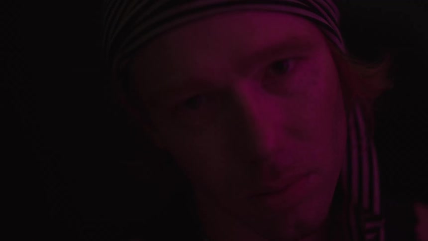 A closeup shot of a man wearing a striped bandana and a tank top with his face illuminated by a pink light in a dark setting