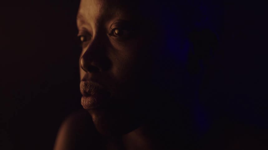 A close view shows a woman with dark skin in profile with her head turning to look over her left shoulder while a warm orange light illuminates the left side of her face