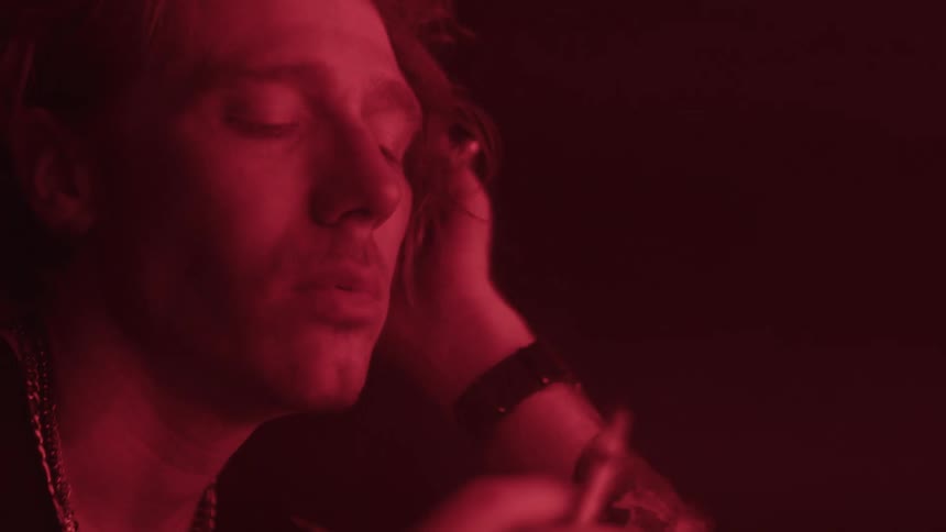 A man with light colored hair and a chain necklace smokes a lit cigarette with his eyes closed and his face partially illuminated by a deep red light