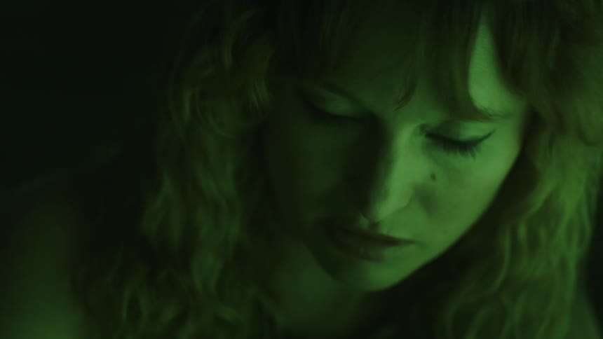 A woman with wavy hair gazes upward with an intense serious expression in dim green lighting