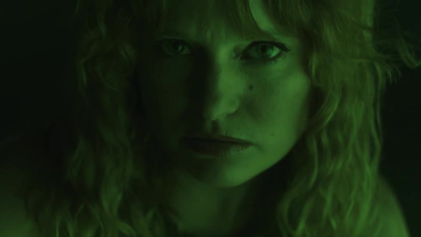 A womans face with a serious and intense expression dramatically lit by a green light source in an otherwise dark and abstract environment