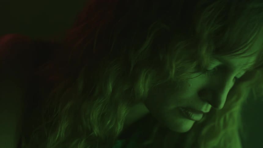 A womans face illuminated by a dramatic green light as she lifts her head to look intensely into the camera