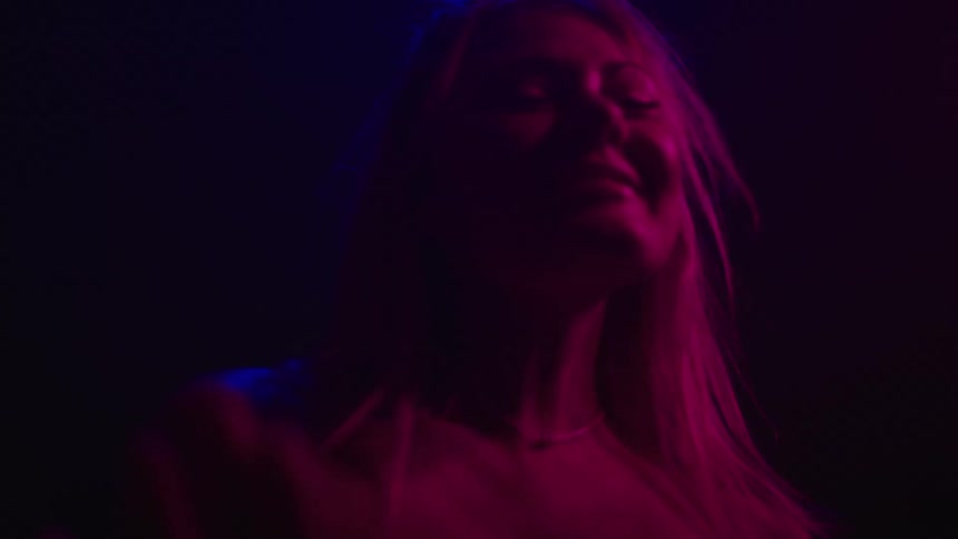 The video shows a woman with blonde hair dancing in a dark room under pink and blue lighting she is smiling and appears to be enjoying herself