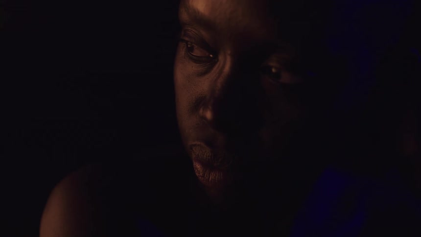 A young black woman shirtless in a dark room looks restless as she moves her neck back and forth