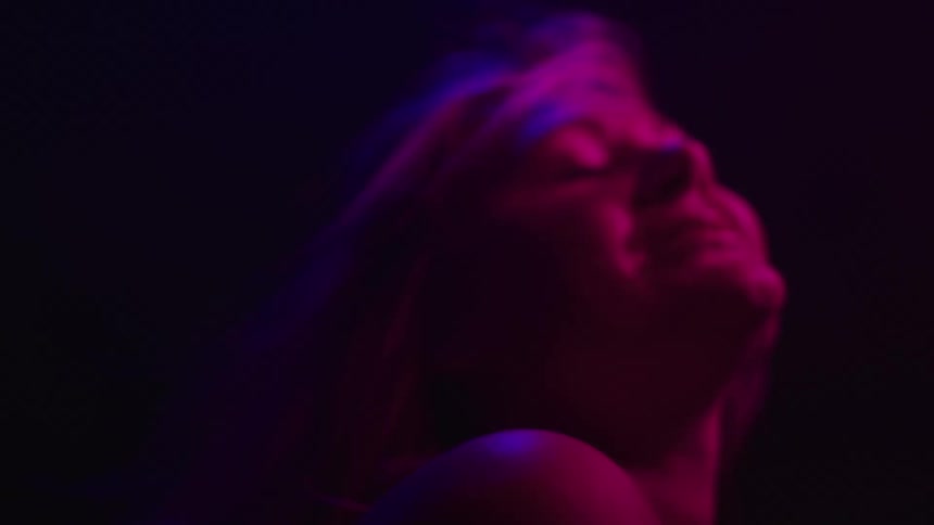 A woman smiles and flips her hair in dramatic pink and purple light