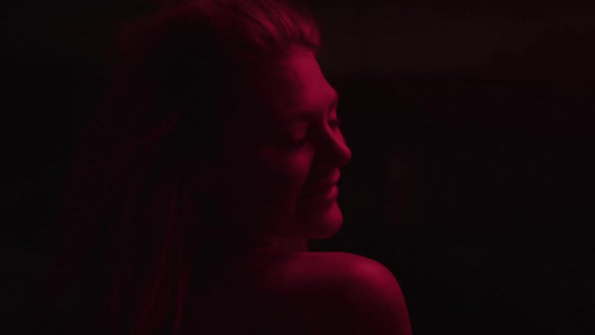 A woman in red lighting flips her hair dramatically while turned away from the camera