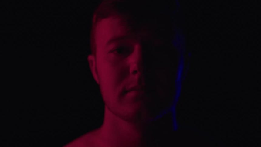 The video shows a mans face in a dark room his face is lit from the side by both red and blue light casting a strong shadow on the opposite side of his face