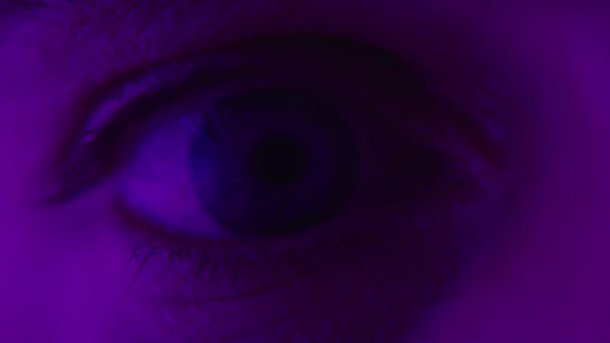The video shows a dark scene and then an extreme closeup of a persons eye is illuminated with a pulsing pink light the scene then goes dark again