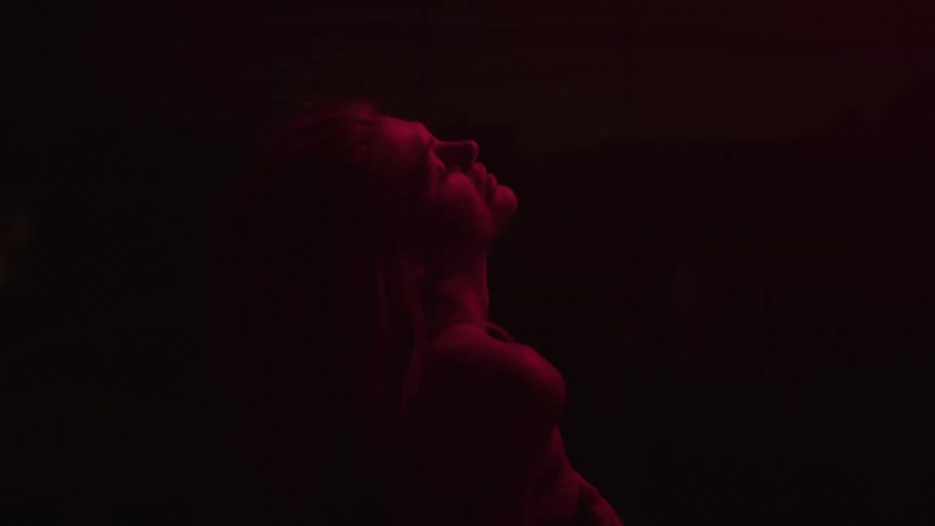 A video shows a woman with blonde hair standing in a dark room with a red light illuminating her she appears to be dancing slowly raising her head and hands as she moves to the music