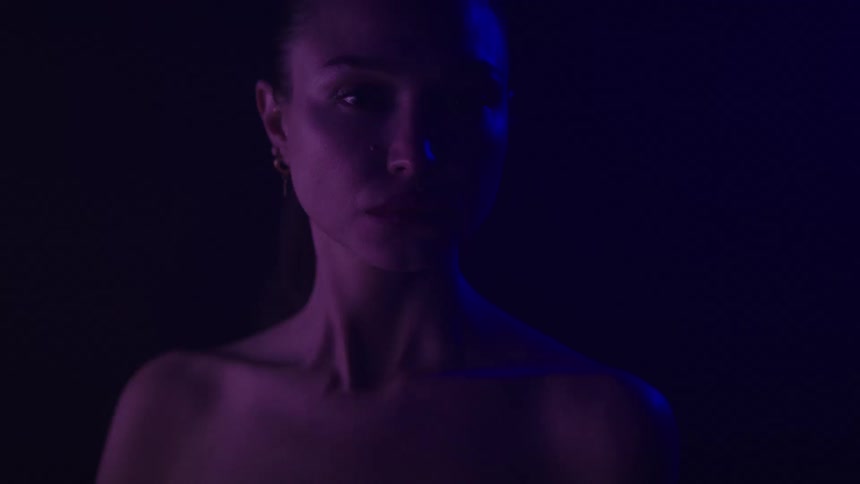 A woman with her hair pulled back with a blue light illuminating her face from the side as she slowly turns her head to face the camera with a serious expression