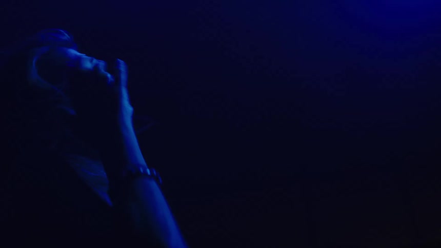 A woman with light colored hair and a thin chain necklace is seen in silhouette with a blue light illuminating her face and hair as she tilts her head back and forth