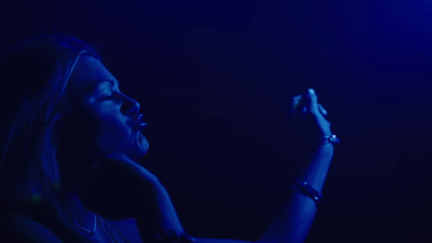The video shows a woman with blonde hair wearing a white top and a bracelet singing into a microphone the setting is dark with a strong blue light