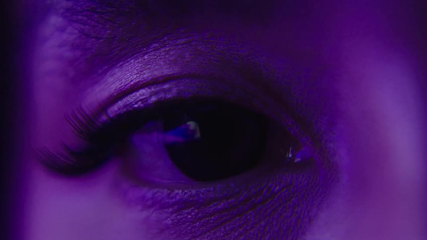 A close view shows a persons eye with a dark iris and long eyelashes as the light from a flashing screen is reflected in their eye