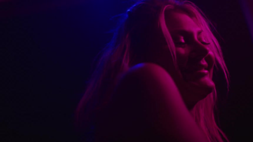 A close view of a woman with long light colored hair shows her tilting her head back then smiling as a bright magenta light illuminates her from the right and a blue light illuminates her from the left