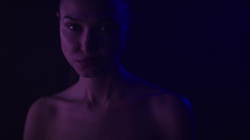 A woman with bare shoulders is illuminated by soft purple lighting gazing forward with a calm neutral expression