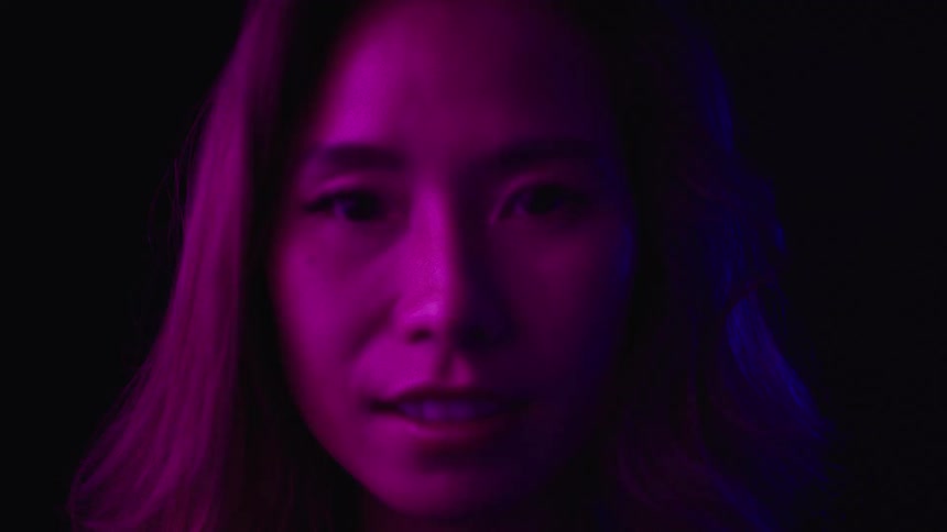 A young asian woman with long hair stands in a dark room looking around as disco lights reflect on her face