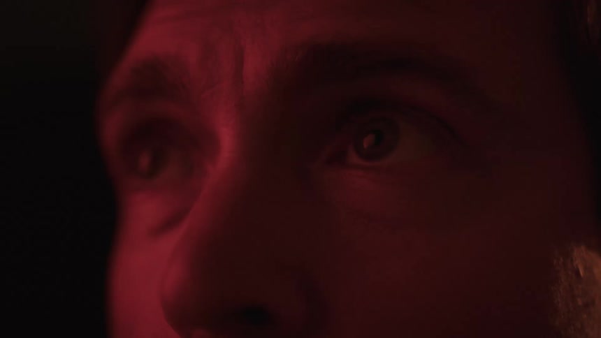 A closeup of a mans face in the dark with his eyes wide open as a red light illuminates his face from the side