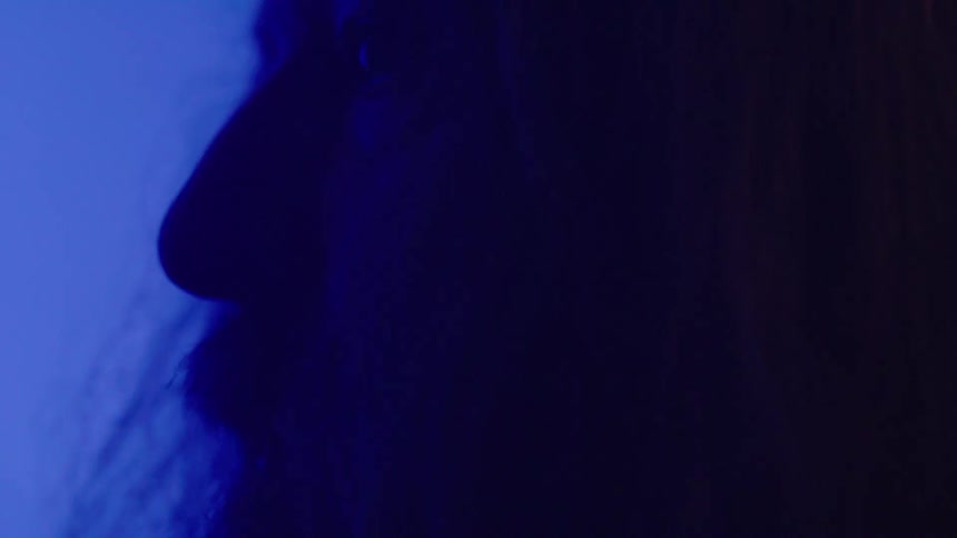 A bearded man sits inside a dark room illuminated by dark blue light at night