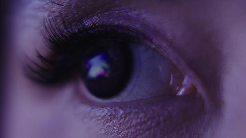 A woman blinks repeatedly her long lashes catching the light reflected from a screen on her face