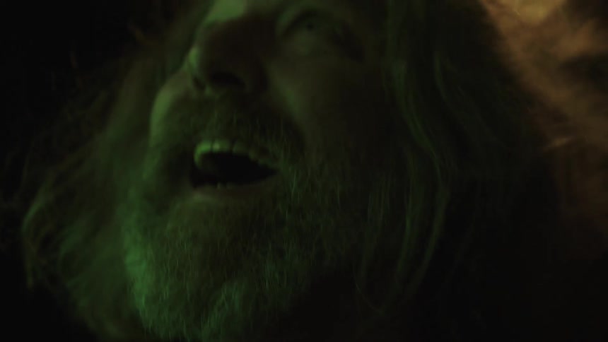 The video shows a man with a long graybrown beard and long hair looking offcamera with a surprised or wideeyed expression he is illuminated by a green light and his mouth is slightly open as if in the middle of speaking