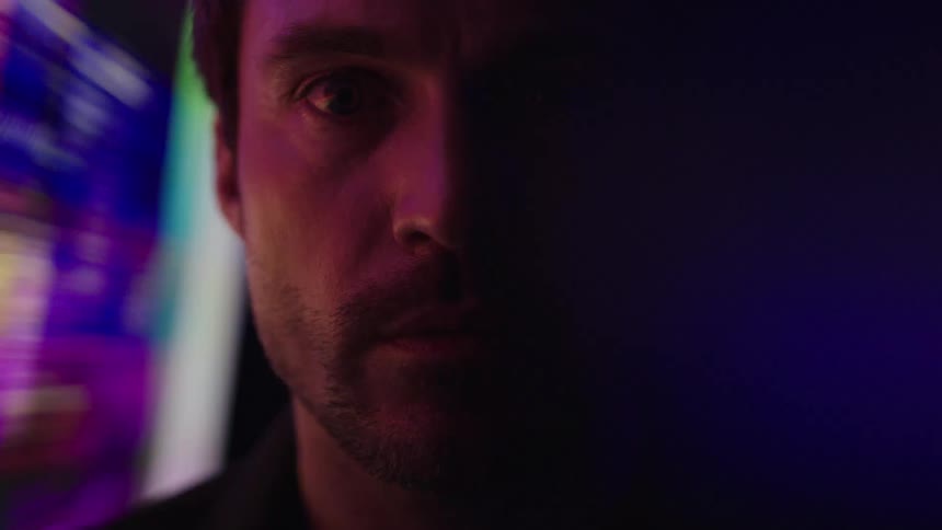 A mans face is seen in a close view with his eyes looking directly into the camera as a colorful flashing screen illuminates the left side of his face