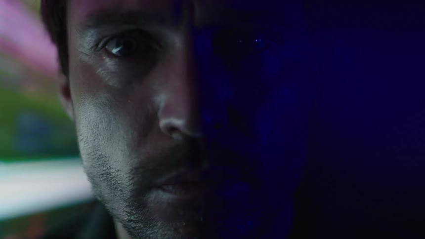 A mans face is seen in a close view with his eyes looking directly into the camera as a colorful flashing light illuminates the left side of his face