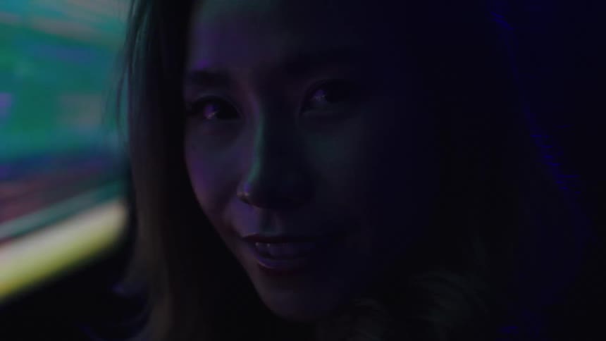 A young asian womans face is illuminated by green and pink lights as she smiles directly at the camera