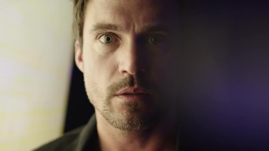A mans face is seen in a close view with his eyes looking directly into the camera as a colorful flashing screen illuminates the left side of his face