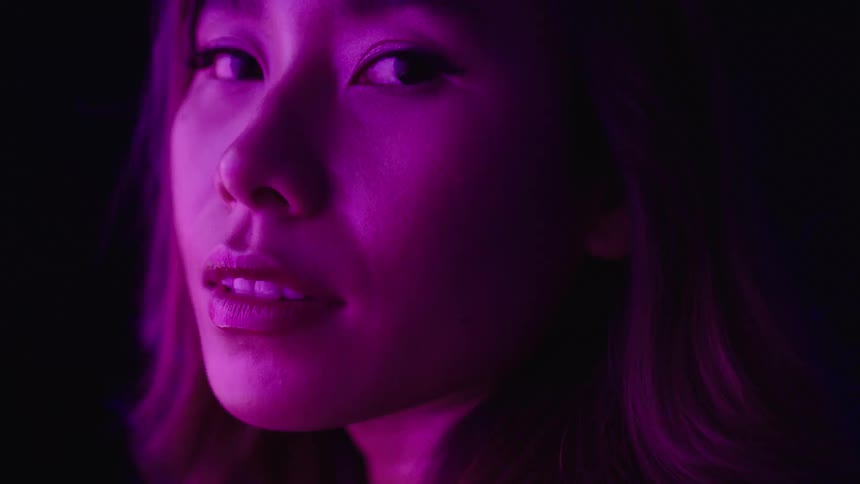 The video shows a persons face from the side with the features obscured by deep shadow a faint blue and purple light flashes over them highlighting their profile