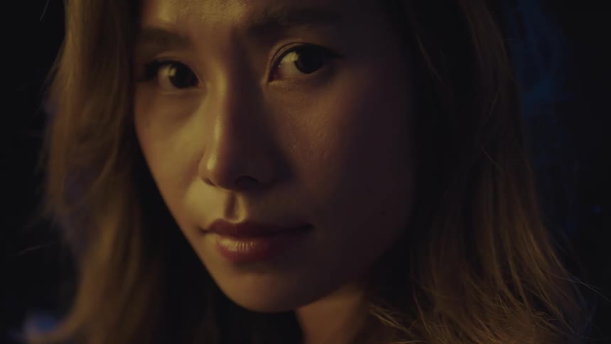The video shows a young woman with long blonde hair and a serious expression lit from the side by a light source she slowly turns her head to face the camera maintaining a steady gaze