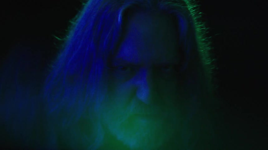 An angry looking mans face is lit with green and blue light in darkness