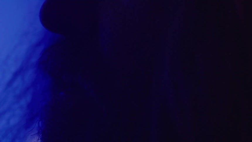 A middleaged man with long hair and a long beard stands in a dark room illuminated by dim blue light
