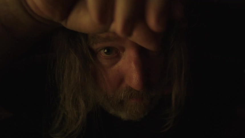 Bearded man with long hair stares directly forward in a dark setting
