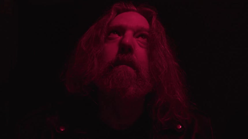 A man with a full beard and long hair looks directly up while a red light shines on his face and hair