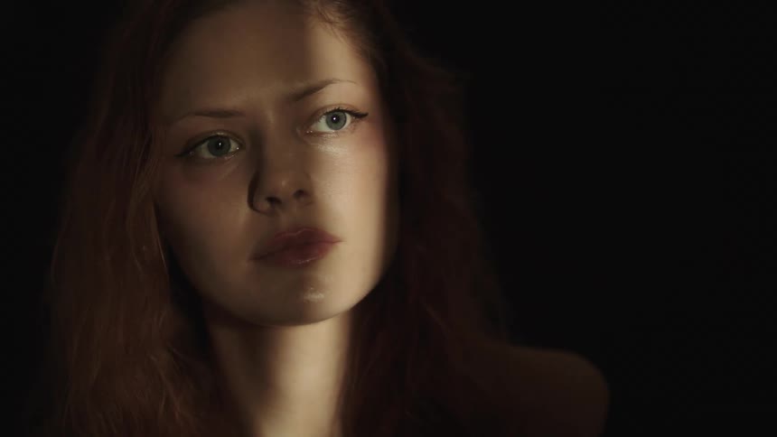 A close view shows a woman with long light brown hair and a serious expression looking directly into the camera while the left half of her face is illuminated by a beam of warm light