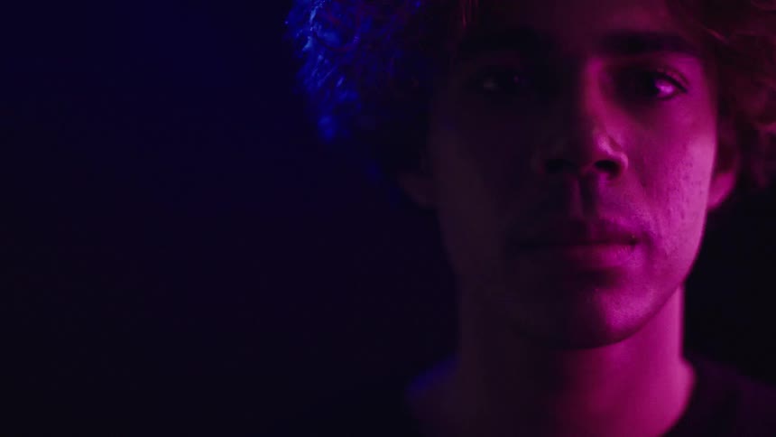 A closeup shot focuses on a mans face in low light with his features illuminated by a colorchanging light from the left side
