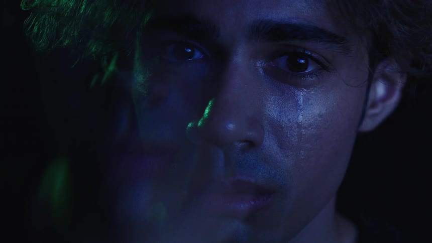 A sad mans face partially illuminated by colorful blue and green lights is shown in a closeup shot