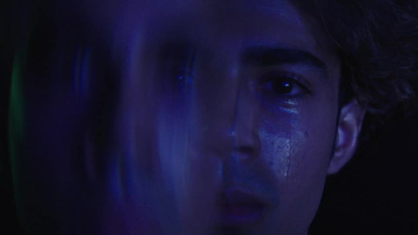 A young man sits in a dark room with faint light reflecting on him revealing a face filled with sadness and tears in his eyes