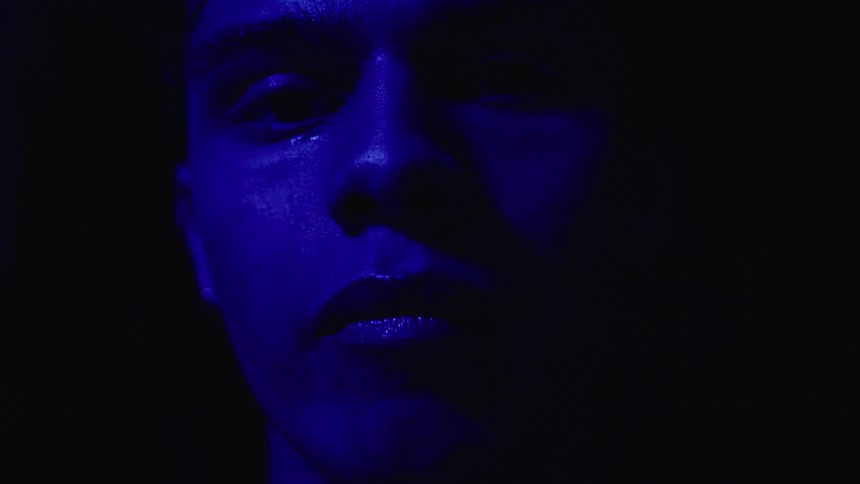 A close view of a mans face in a dark space with a bright blue light illuminating the right side of his face as a tear falls from his eye